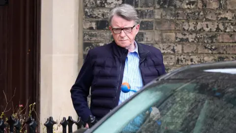 Mandelson looks down as he exits the front door of his home holding a dog toy in front of a car 