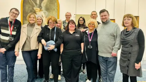A group of people have are stood smiling whilst looking at the camera. They are in an art gallery, with pictures on a light-coloured wall behind them.