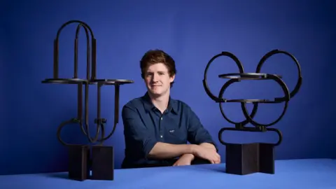 Cameron Pearson pictured sitting with his arms crossed at a blue table with a blue background next to two of his metal sculptures. He has brown hair and is wearing a dark blue button shirt. 