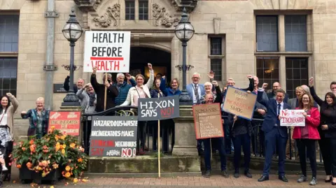 A group of people gathered outside the front entrance of a town hall. Some are holding placards with slogans written on. Many are holding their hands in the air.