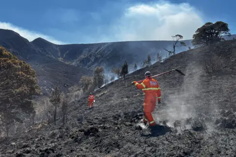 RSPB Scotland Two people wearing bright orange overalls walk across a scorched, smoking hillside.