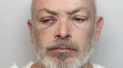 Police photo of Steven Mahon who is looking downwards. He has a cut under one eye and has a grey beard. He is wearing a white T-shirt.