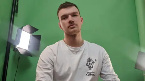 John O'Reilly John O'Reilly looking at the camera with a green screen behind in the background, wearing a white top and necklace.