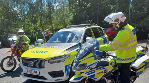 A blue, white and yellow police car is parked and there are three police officers next to it. On the right there is a policeman in a yellow fluorescent jacket and white helmet. He is standing next to a yellow, white and blue police motorbike. On the left are two police officers. One is wearing all black police uniform and is getting into the driver's side door which is open. Next to him is another police officer who is holding a red and yellow off-road bike. The officer is wearing yellow fluorescent gear and white helmet