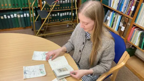 Victoria Evans A photo taken above archivist Victoria Evans, dressed in a brown jumper and black and white gingham skirt, sat at a table, turning the pages of one of the tiny books lying in front of her on a wooden table. They are all hand-illustrated and handwritten in what looks like pencil. Behind her are bookshelves loaded with books and folders.