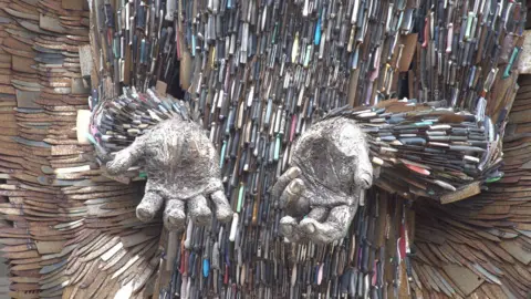 A close up of the Knife Angel's front body and hands. Knives and knife handles are clearly visible. The handles of the knives all seem to be different colours. The wide blades are rusting.