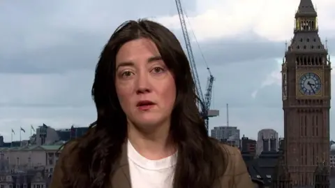 Sarah Owen, with long dark hair, is pictured in a television studio interview setting, wearing a light-coloured top and checked blazer. Behind the person is a view of central London, including the Elizabeth Tower (Big Ben), city buildings and a construction crane beneath a cloudy sky.