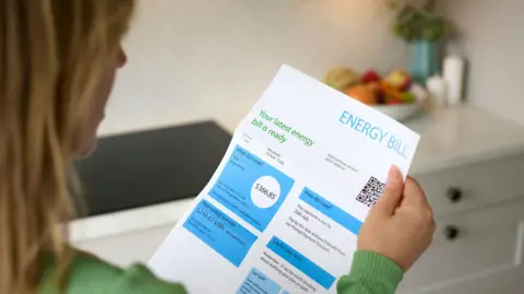 Over the shoulder of a woman looking at an energy bill in her kitchen with a fruit bowl in the background.