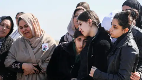 Reuters feminino lamentam um funeral em Latakia, vestindo vestes cinza e preto e algumas escorburas na cabeça, chorando e confortando uma à outra