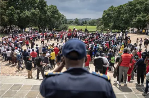 AFP Man security guard standing in a dark blazer and hat in front of a crowd of students.