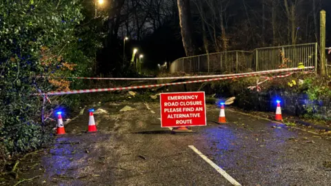 A red sign saying emergency road closure in the middle of a road which is taped off by red and white striped tape and there are bushes on either side. Behind the sign appear to be branches and bits of a tree.