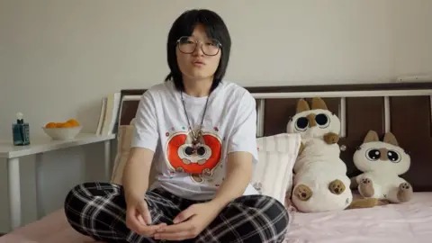 Enxu sitting cross-legged on a bed, with two soft toys next to her. She is wearing large glasses, and has black, neck-length hair, cut with a fringe. She is wearing a white T-shirt with a red image on it, and checked trousers.