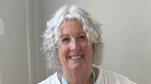 Bradford Teaching Hospitals NHS Trust A woman with curly white hair smiles at the camera. She is wearing a white shirt and blue lanyard