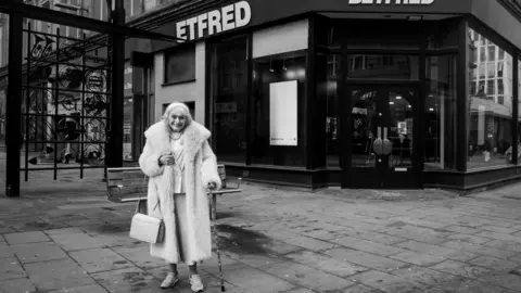 John Bolloten A black and white photo of an elderly woman wearing a fur coat stood outside a betting shop in Bradford city centre.