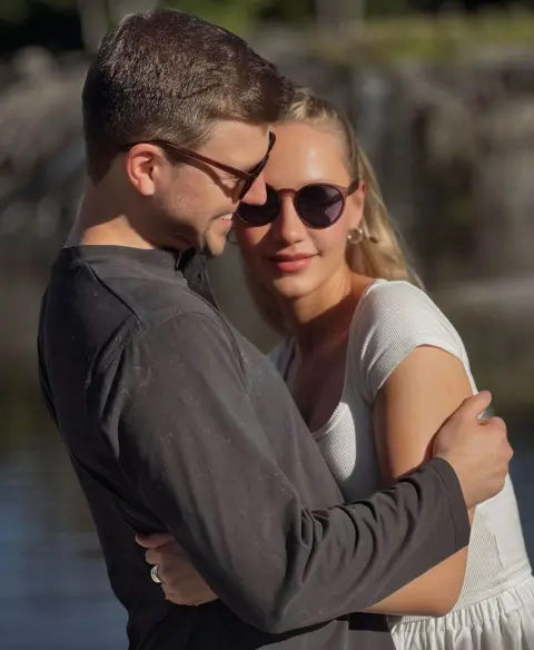 Handout Two people standing close together in the foreground, with arms around each other beside a calm body of water. One wears a dark long‑sleeved top and the other a light short‑sleeved dress. Behind them is the water, trees and greenery.