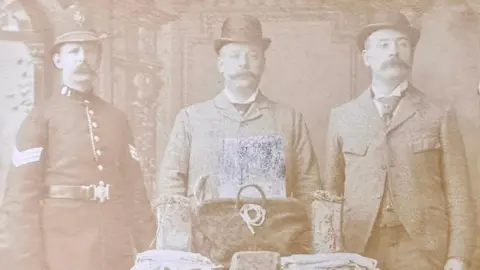 Thames Valley Police Museum This is a black and white sepia-style posed photograph of three men, all wearing large moustaches which were fashionable in the late nineteenth century. The man on the left is dressed in a police uniform while the two men to his right are wearing Victorian suits and ties. Among the evidence on a table in front of them is a bag pulled out of the river, the brick it was weighed down with and some fabric. 