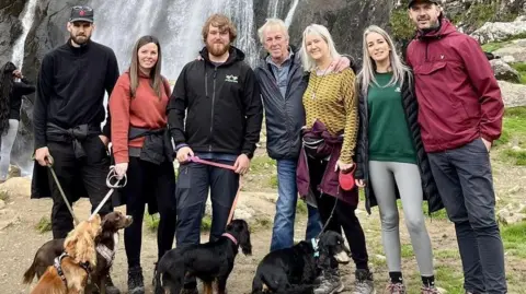 Brain Tumour Research A row of seven people in front of a waterfall.  Four are holding dogs on leads. Alex with short hair and beard and Trevor with short white hair are in the group.