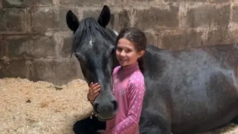 Nenthorn Equestrian Centre A girl of about 11 years is kneeling beside a black horse that is lying down. The horse's head is cradled in her hands and she is smiling. They are in a stable.