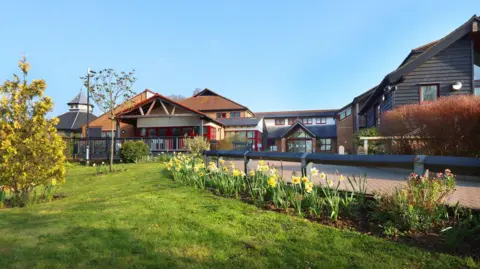 Farleigh Hospice A modern two-storey building in the background, with a paved road or walkway leading up to it. There are daffodils in the foreground and a grassy lawn. There are blue skies above.