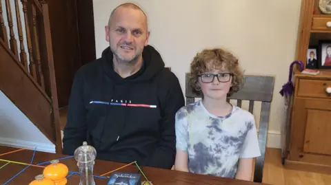 Richard Clark Harry is looking at the camera. He has black rimmed glasses and curly blonde hair. Richard sits beside him with a black hoodie and a blue, white and red line across the middle. They are sitting at a wooden table in a kitchen with a dresser behind. 