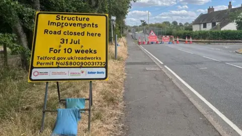 Ellen Knight/BBC Photograph of a yellow sign, reading in black letters: "Structure Improvements, road closed here, 31 Jul, for 10 weeks." It is placed on a grass verge next to a road. Beyond the yellow sign are grey metal fences and orange traffic cones blocking off the bridge. Red signs with "road closed" on them in white letters are placed in front of the cones.