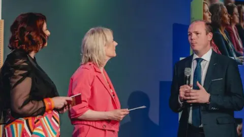 Three people on a stage. A woman on the left has shoulder length red hair. She is looking away from the camera. She is wearing a black mesh shirt and colourful stripy trousers. In the middle a woman with blonde hair is looking at the minister. She is wearing a pink matching suit. On the right, the minister is wearing a grey suit and a blue tie. He has short brown hair and is holding a microphone. 