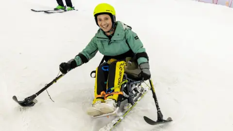 Cameron Ross Hall/Holmlands Ellie skiing wearing a yellow helmet, green ski jacket, yellow and black trousers and white trainers. She holds two ski poles out to either side and has a big smile