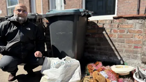 Gabby Colenso/BBC Naveed Shakoor has a grey beard and glasses and is looking at the camera. He is wearing a black North Face jacket and is squatting beside a bin with some dumped rubbish beside it, in front of a brick, terraced house.