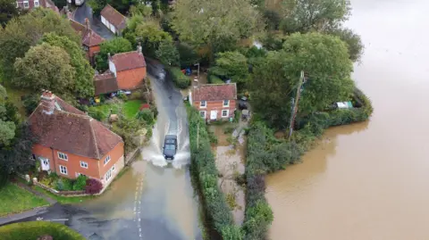 An aerial view showing a car driving through a flooded road, with water spraying out either side of it. Around the road there are red brick houses with flooded gardens and a flooded field can also been seen.