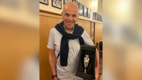 Tony Jones Tony Jones wearing a pale shirt with a dark jumper draped over his shoulders. He is smiling and holding a box containing a small silver trophy.