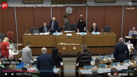 Oldham Council/YouTube Councillors stand up to leave. The mayor is at the centre of the desk with a gold chain around his neck and shoulders.