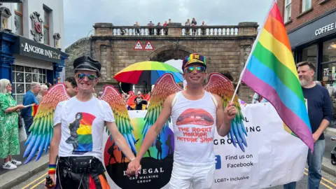 Two men stand, smiling at the camera. They are holding hands, and one of the men has a rianbow flag. The man with the rainbow flag is wearing a white top which has the words 'THE ROCKY HORROR PICTURE SHOW' written on the front. He is also wearing sunglasses and a rainbow-coloured hat. The man next to him is also wearing sunglasses, as well as a black hat and a white t-shirt. Both men have rainbow-coloured angel wings on their backs. 
