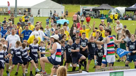 Sarah Elisabeth Photography/Wooden Spoon A large group made up of dozens of children, and adults, mostly wearing rugby kit, are performing warm-up exercises on a field. In the background tents and gazebos can be seen, with more people walking along them.