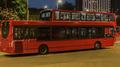 A single-decker red bus, with a double-decker behind it.