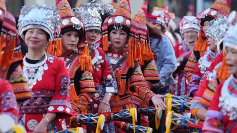 PA Media A group of women wearing colourful, traditional Asian clothing, including silver headdresses and red embroidered outfits, stand in a row holding decorated drumsticks.