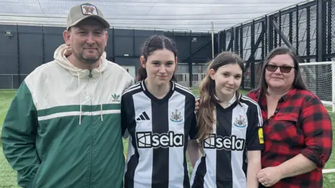 David Dalton, Holl Dalton, Olivia Dalton and Sherrie Dalton stand on an astro turf football pitch. Mr Dalton is wearing a cream and green Adidas sports jacket and a Liverpool football cap. Holly and her sister are wearing Newcastle tops. Both girls have long dark hair. Sherrie Dalton is wearing a red and black checked flannel shirt. They are standing close together and smiling. 