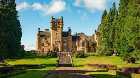 BBC/ Studio Lambert A sandstone castle with a turret, and stairs leading down to a large garden with grass an trees at either side.