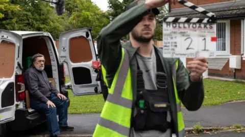 As fotos de limão Ricky Tomlinson, no personagem de Bobby Grant, estão vestindo jeans e uma camiseta cinza. Ele está sentado na parte de trás de uma van em uma rua residencial conhecida como Brookside Close.