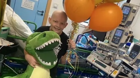 GOSH Harley, who is wearing a black T-shirt, holds a large teddy dinosaur and balloons. He has a shaved head due to his chemotherapy and is standing in front of hospital equipment and technology. He is smiling.