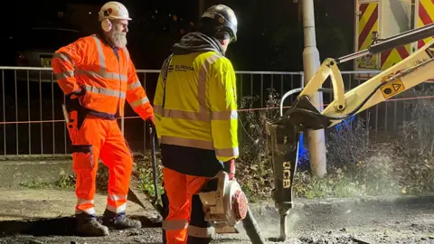 Road resurfacing in Laindon High Road. Two men in orange overalls watch a JCB drilling arm break up a road surface.