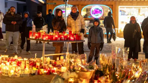 People look at tributes and candles left for victims of the fire at Swiss ski resort. It is nightr time and there is a glow from the candles