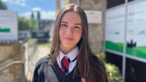 Teenager Molly with long brown hair smiles to the camera. She's wearing school uniform including a black and red tie.
