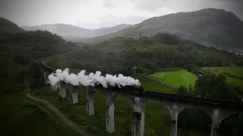 A steam train going over a viaduct with a plume of steam trailing behind and hills in the background 