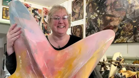 A woman smiles as she stands holding a large painted starfish sculpture in a gallery full of paintings. She has blonde hair pulled back and wears glasses and a black top. The starfish is painted in pinks, peaches and blues.