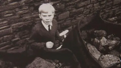 Getty Images black and white picture of Jeff Edwards aged eight, with blonde hair, a dark jumper, white shirt and dark tie, holding a toy plane, looking at the camera, a few days after being rescued 