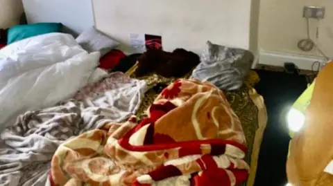 A sparsely furnished room with bedding spread directly on the floor. Several blankets and sheets are piled on patterned floor coverings beneath a chimney breast or wall recess marked by dark damp or mould stains. 