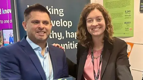 Sarah Coombes A man and woman stand next to each other in front of a billboard. Both are smiling. He has black hair and is wearing a navy suit jacket and blue shirt. She has curly brown hair and is wearing a pink blouse and dark jacket and has her hand on his shoulder.