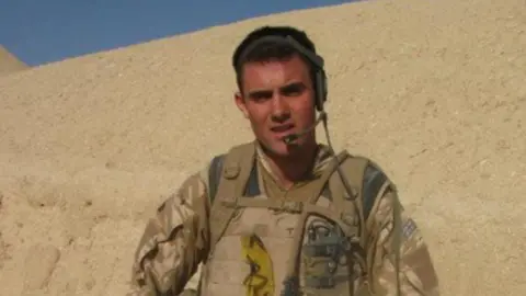 Rhys Thurtell looks directly at the camera as he stands on a sandbank. He is wearing a headset and an Army uniform. 