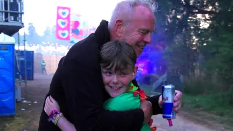 A man with grey hair and black jacket hugs a young boy with brown hair and green T-shirt.