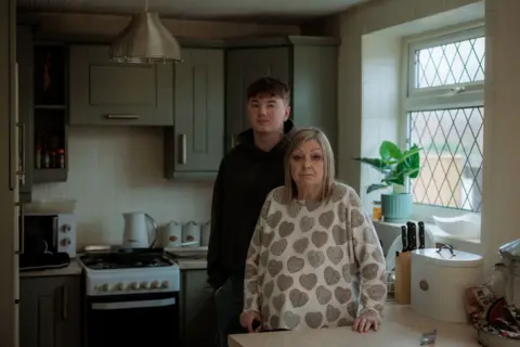 Two people stand at a kitchen counter in the foreground, one Alex's nan Susan is wearing a patterned jumper and resting hands on the worktop, Alex is in a dark hoodie positioned slightly behind. On the right, a countertop holds a wooden knife block, a white container with lid, and a bowl near a window with leaded glass panes and a potted plant. In the background, a cooker with hob, kettle, toaster and spice jars sits beneath grey cabinets, with a metal pendant light overhead.